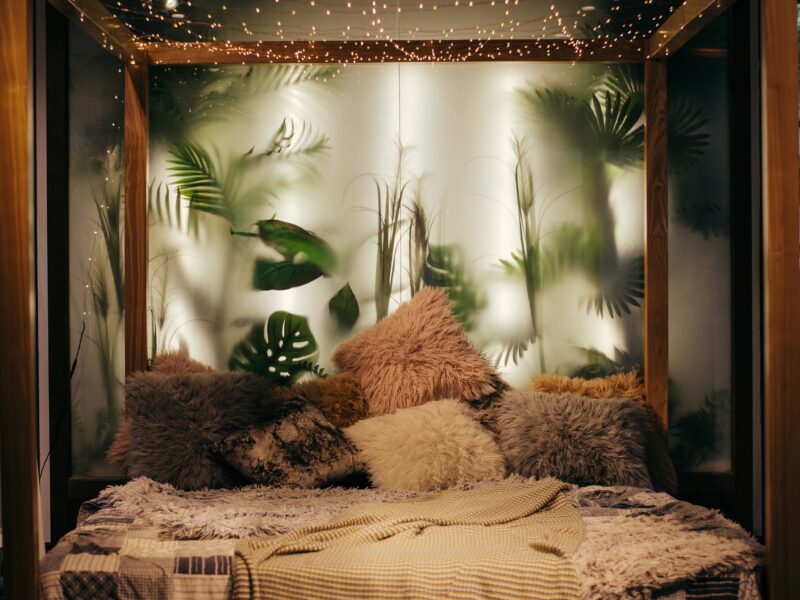 Image of a sensory safe space with bed frame, string lights, fuzzy pillows, soft blankets, and back-lit glass behind the bed with plants