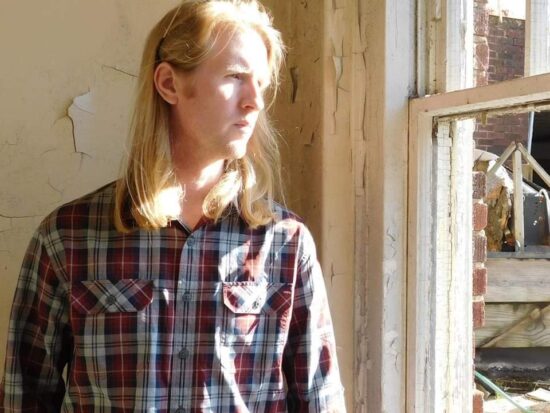 Man with shoulder-length blonde hair look stands next to a window looking outside. Image is connected to a story about autism diagnosis in adulthood.