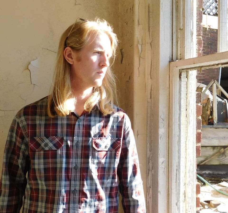 Man with shoulder-length blonde hair look stands next to a window looking outside. Image is connected to a story about autism diagnosis in adulthood.