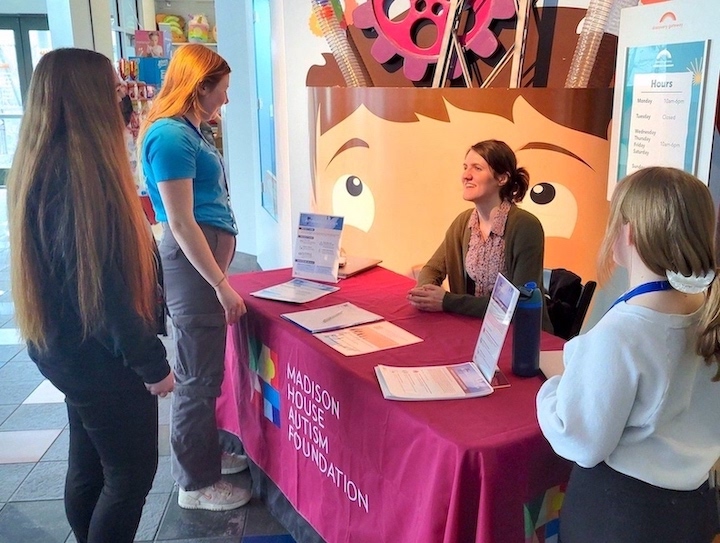 Smiling woman behind table with MHAF logo speaking to three young women. Background shows fun kids images at the children's museum.
