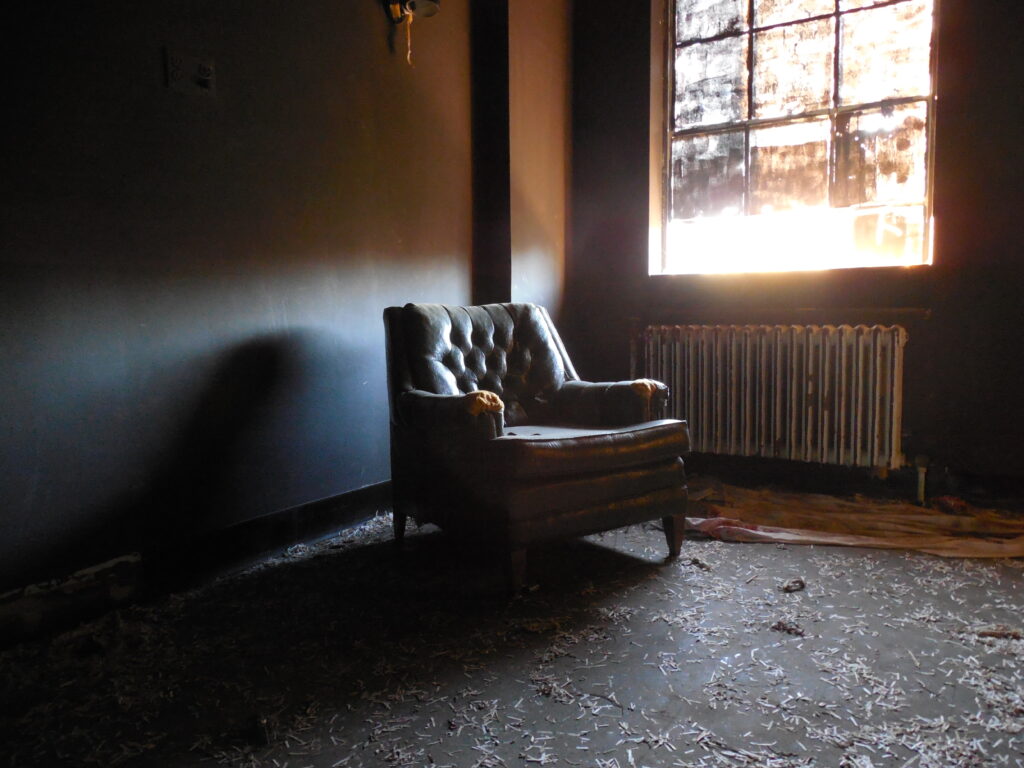 Abandoned room with light shining from a window onto a brown chair. Image is connected to a series of photography images from an autistic artist story about autism diagnosis in adulthood.