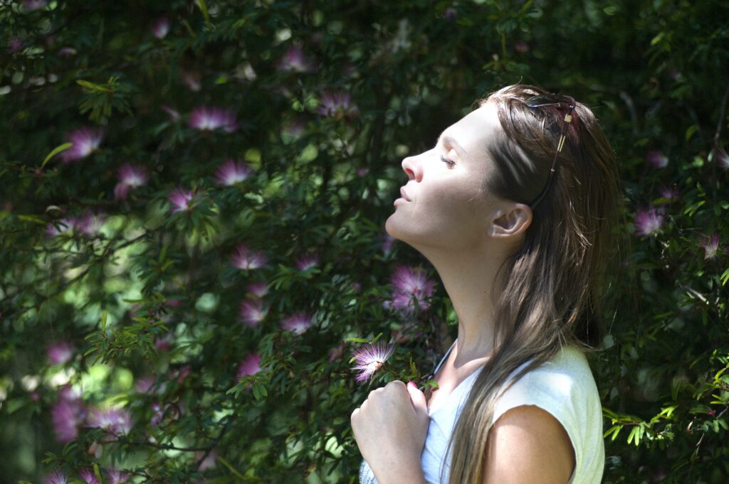 Shows a profile view of a young person with long hair who is outdoors with green plants behind them. They have their eyes closed and their head tilted up slightly with a calm expression on their face. It represents a type of stress management.