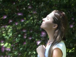 Shows a profile view of a young person with long hair who is outdoors with green plants behind them. They have their eyes closed and their head tilted up slightly with a calm expression on their face.