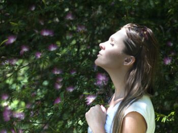 Shows a profile view of a young person with long hair who is outdoors with green plants behind them. They have their eyes closed and their head tilted up slightly with a calm expression on their face.