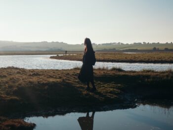Profile view of a woman standing in a patch of grass between two ponds in a watery field with the sun low, staring off into the distance.