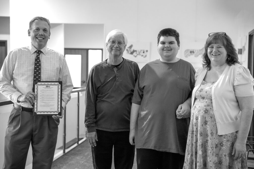 Black and white photo of four individuals smiling at the camera indoors. The mayor of Hutto is on the left holding the proclamation recognizing Autism After 21 Day. The Lyons family is on the right.