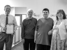 Black and white photo of four individuals smiling at the camera indoors. The mayor of Hutto is on the left holding the proclamation. The Lyons family is on the right.