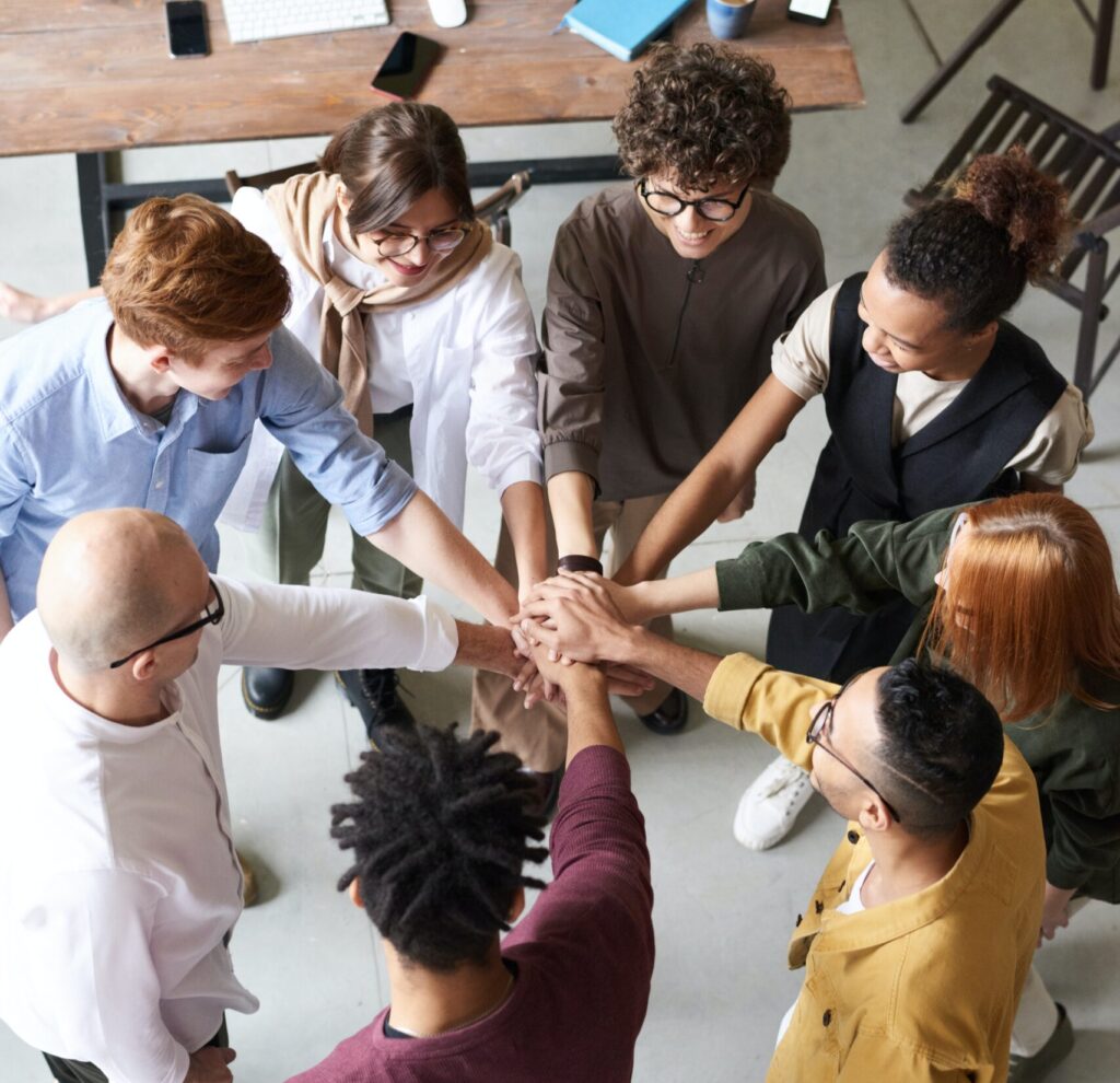 A group of people in a circle each hold a hand out toward the center of the circle in a workplace environment.