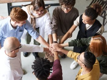 A group of people in a circle each hold a hand out toward the center of the circle in a workplace environment.