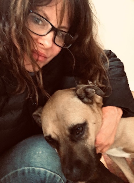 Person with medium length brown wavy hair and glasses is squatting down and cuddling with a short haired dog with creme colored fur with patches of dark brown around the snout and eyes.