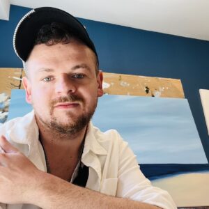 Photo of a person with short hair, beard, and mustache with a baseball cap, white button-up shirt, and paintings in the background.