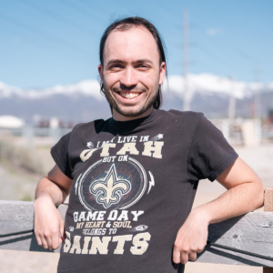 Person smiling outside during the day with a t-shirt on that says, "I may live in Utah but on game day my heart & soul belongs to the Saints". Part of the article about Adam: A Voice for Change.