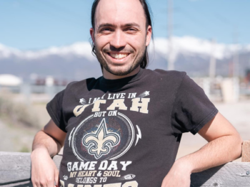 Person smiling outside during the day with a t-shirt on that says, "I may live in Utah but on game day my heart & soul belongs to the Saints"